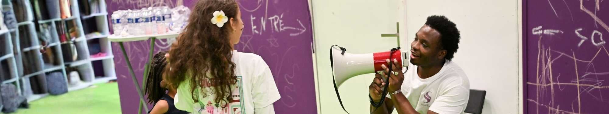 Homme parlant dans un megaphone face a une femme de dos avec fleur dans les cheveux, devant un mur violet griffonne.