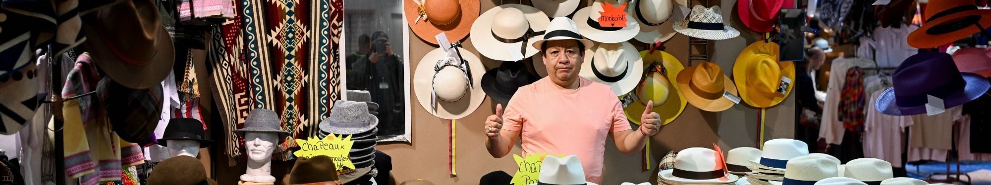 Vendeur sur un stand de chapeaux faisant un signe pouces leves, entoure de nombreux modeles colores exposes au mur.