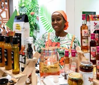 Une exposante souriante, portant un foulard orange et une tenue colorée, se tient derrière un stand présentant des bouteilles et bocaux de produits alimentaires artisanaux.