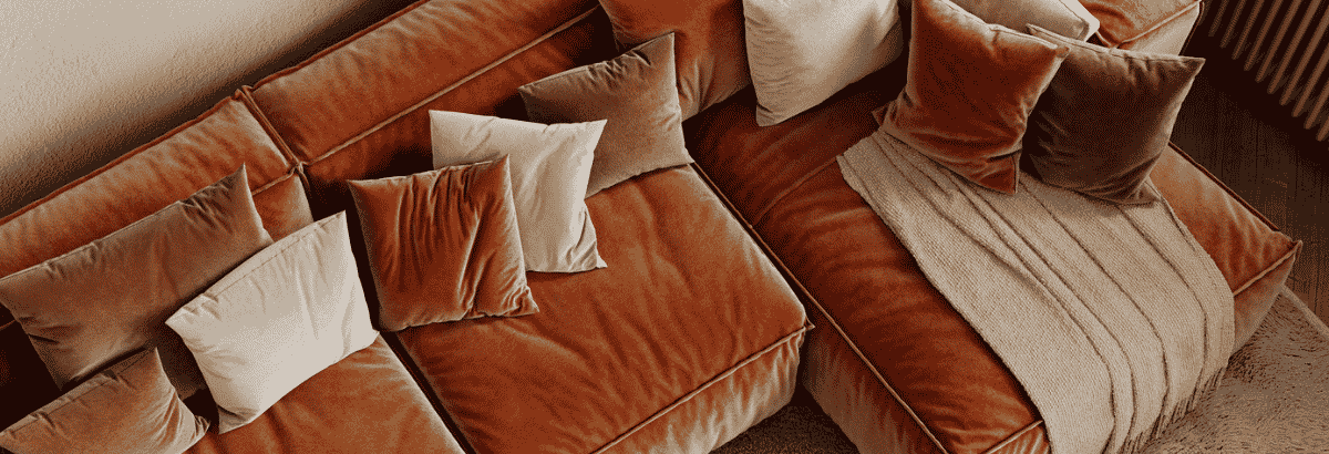 Canapé d'angle en velours brun, vu en plongée, avec plusieurs coussins et un plaid clair disposés sur l'assise.