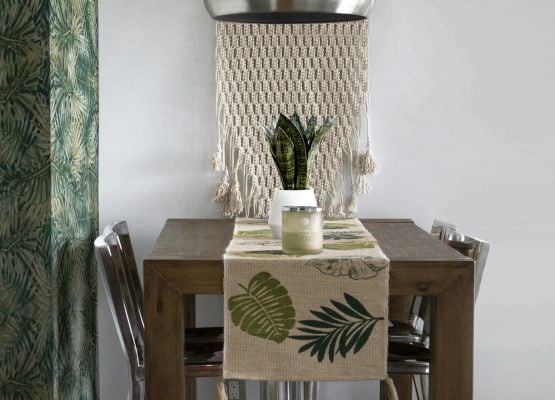 Table à manger en bois avec chemin de table à motifs végétaux, plante en pot et décoration murale textile dans un intérieur épuré