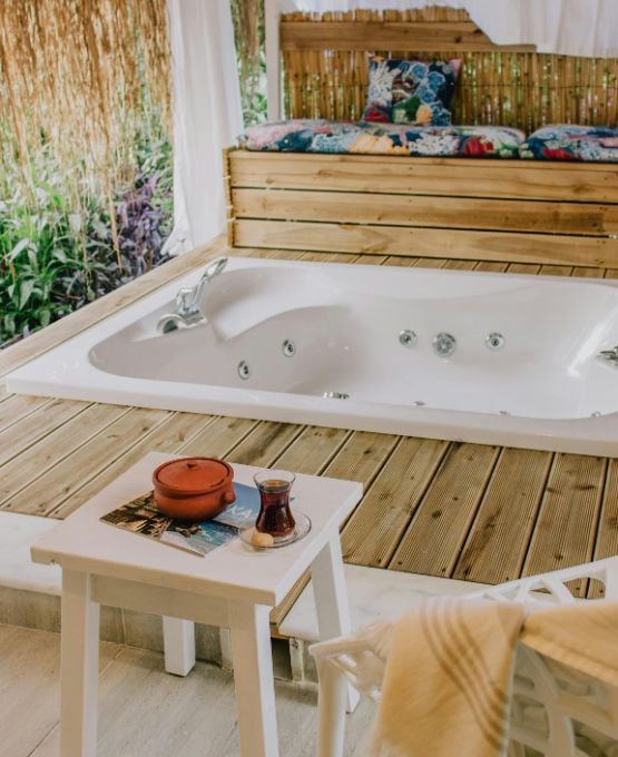Jacuzzi intérieur encastré dans une terrasse en bois avec coussins colorés et table basse avec thé dans un cadre naturel apaisant