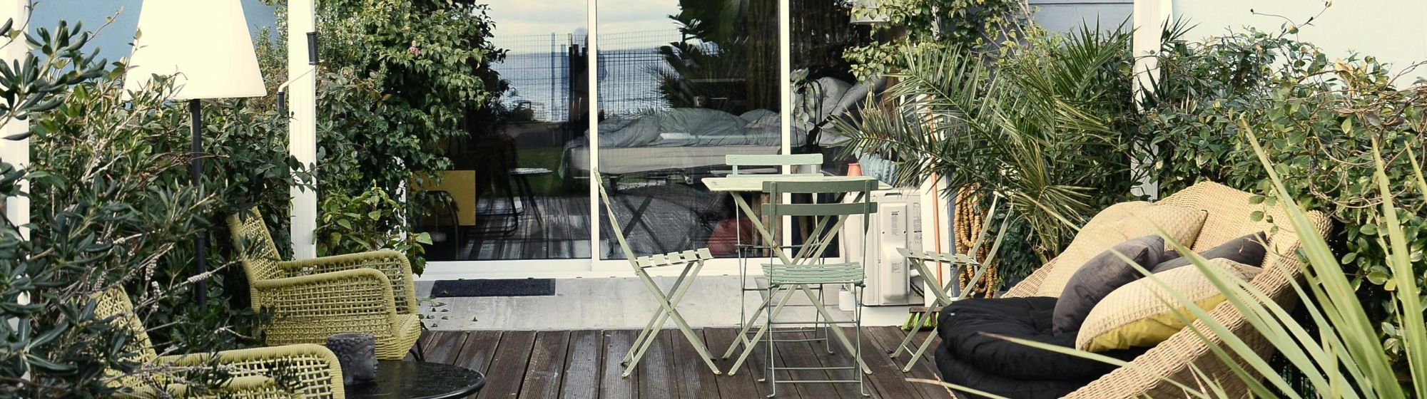 Terrasse extérieure aménagée avec mobilier en bois et métal, entourée de plantes, devant une baie vitrée dans un cadre verdoyant