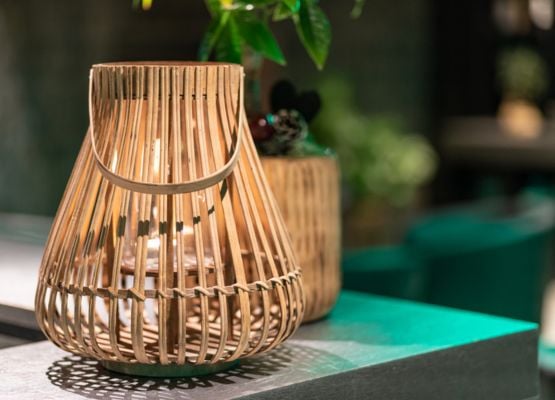 Lanterne en bois tressé avec bougie posée sur une table, avec plante en arrière-plan dans une ambiance chaleureuse