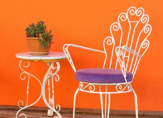 Chaise en fer blanc avec coussin violet à côté d’une petite table ronde avec plante, devant un mur orange vif