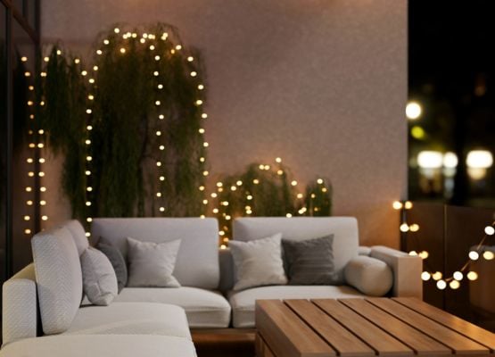 Salon extérieur avec canapé clair, coussins et guirlandes lumineuses devant des plantes, ambiance chaleureuse en soirée