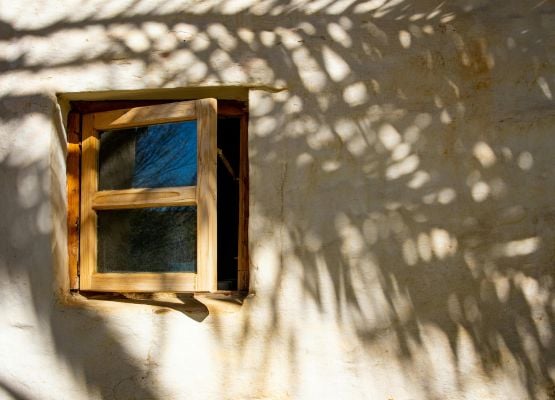 Petite fenêtre en bois ouverte sur un mur clair avec ombres de feuillage projetées.