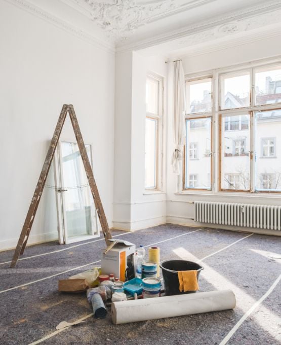 Pièce en rénovation avec matériel de peinture, échelle et grandes fenêtres laissant entrer la lumière.